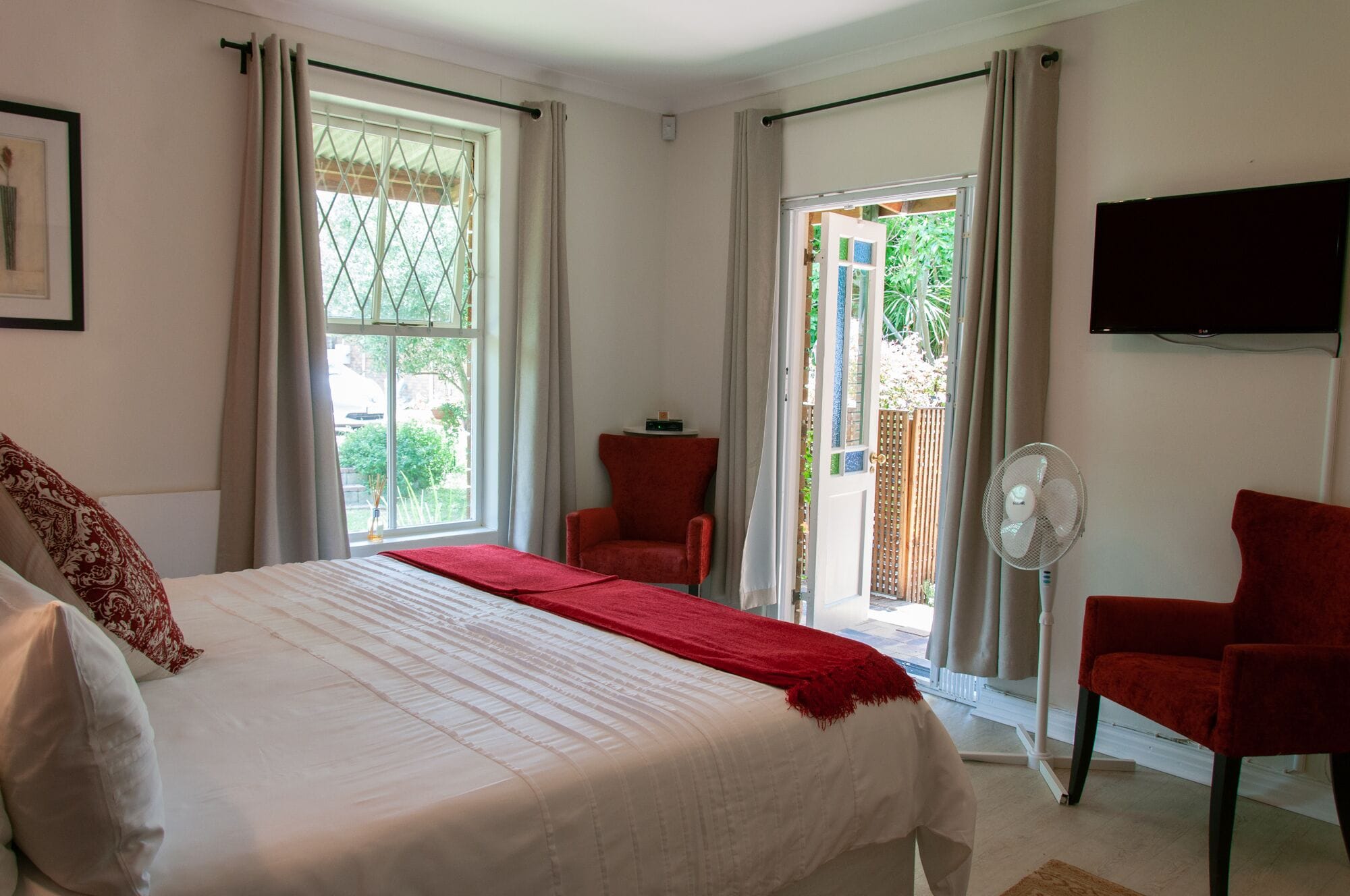 Habitación de hotel con una cama grande, dos sillas rojas, un televisor y una puerta corrediza de vidrio al exterior.
