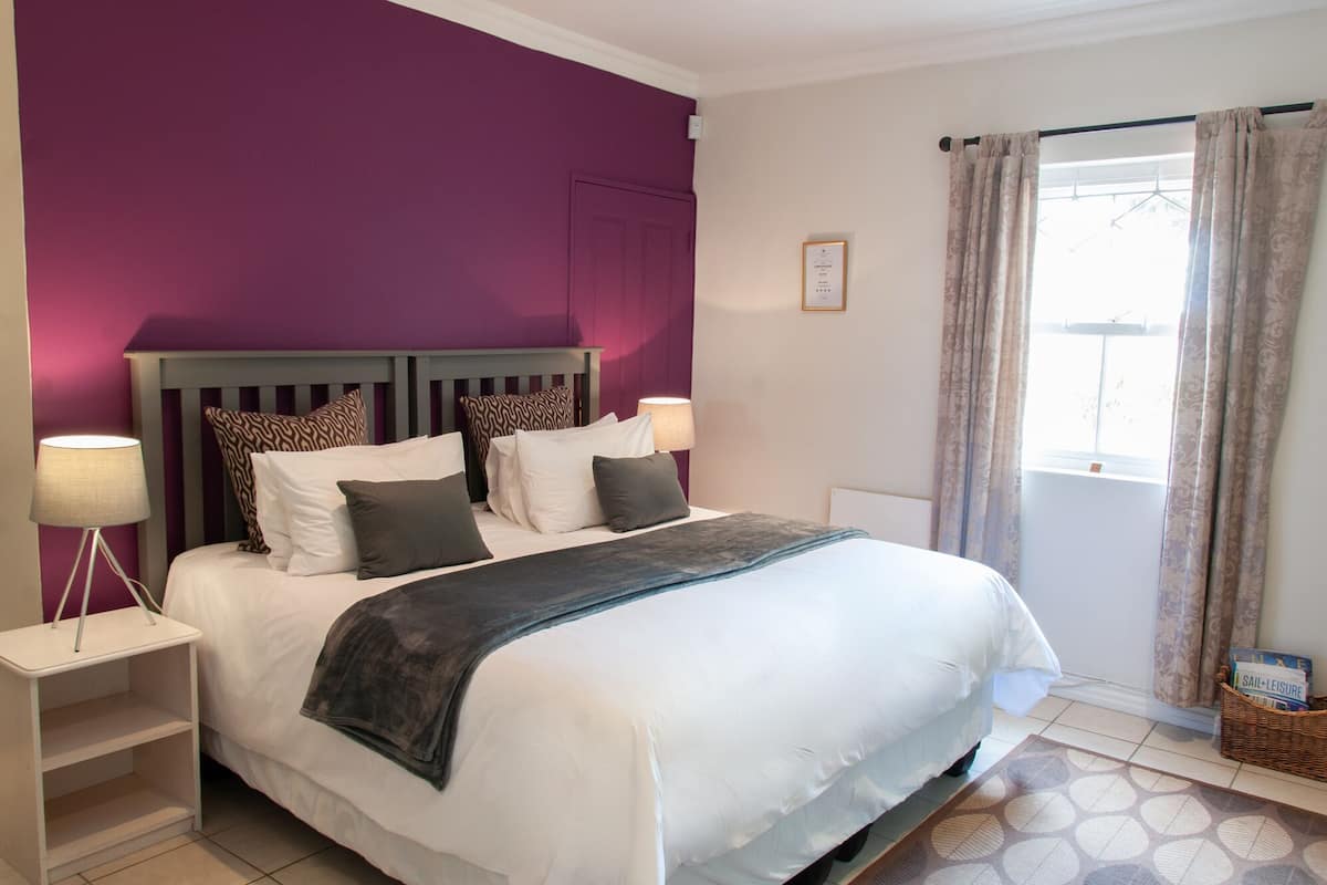 A neatly made bed with a headboard, two bedside tables with lamps, a window with curtains, and a small basket on the floor.