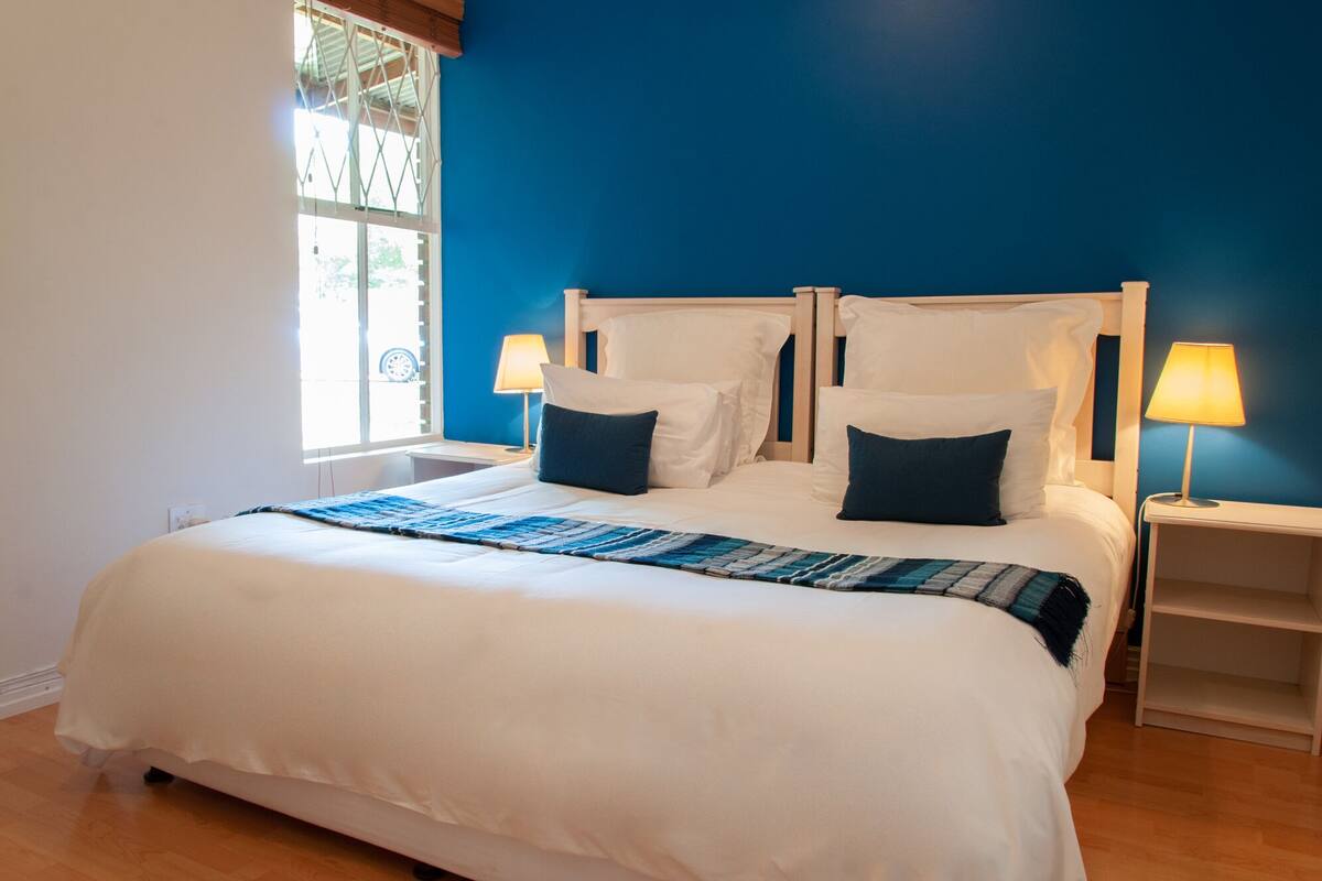 A bedroom with a large bed, two bedside lamps, a window with blinds, and a blue wall.