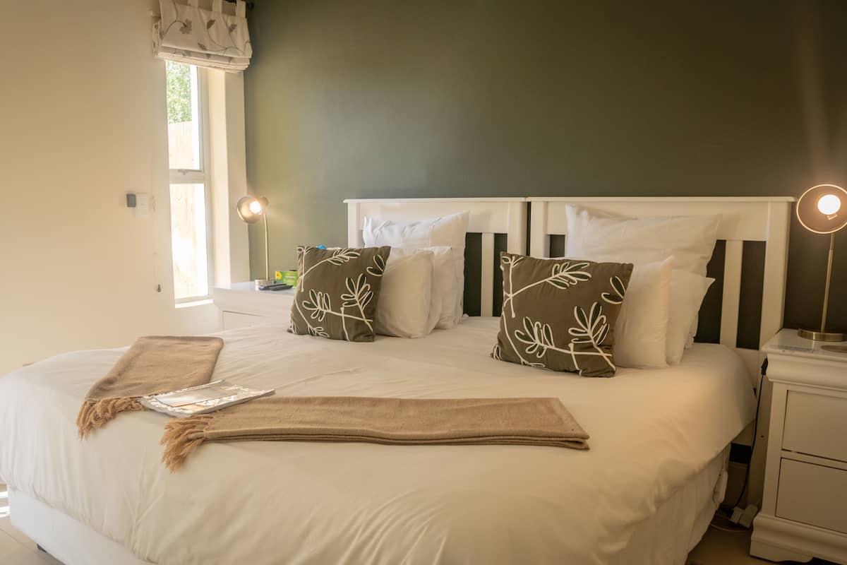 A neatly made bed with white linens and decorative pillows, a bedside table with a lamp, and a window with curtains.