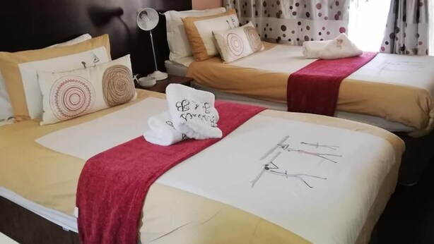 A hotel room with two beds, each with a red throw blanket and a towel with text. - Flossie’s Bed and Breakfast (Soweto)