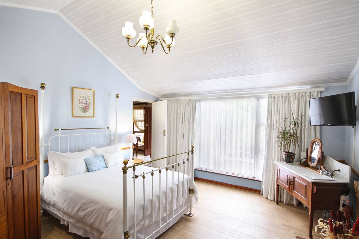 A bedroom with a bed, a wooden wardrobe, a desk with a mirror, and a television.