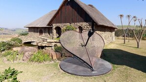 A rustic stone building with a thatched roof, a circular stone structure, and a metal sculpture in the foreground. - Rain Farm Game Lodge (Ballito)
