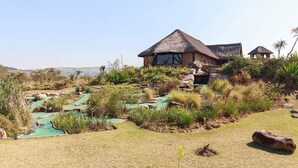 A thatched-roof building with a large window, surrounded by a landscaped garden with pathways and greenery. - Rain Farm Game Lodge (Ballito)