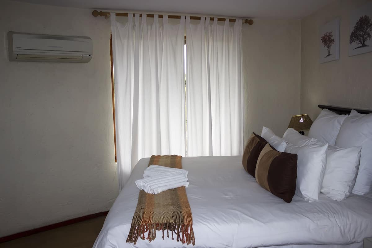 A hotel room with a bed, white curtains, a wall-mounted air conditioner, and two framed pictures on the wall.