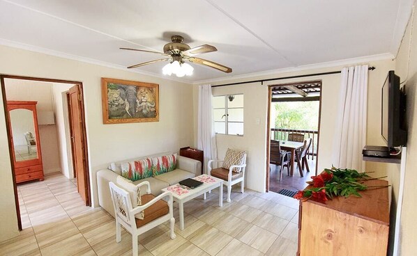 A room with a sofa, two chairs, a coffee table, a ceiling fan, a painting, a wooden cabinet, and a television. - Rain Farm Game Lodge (Ballito)