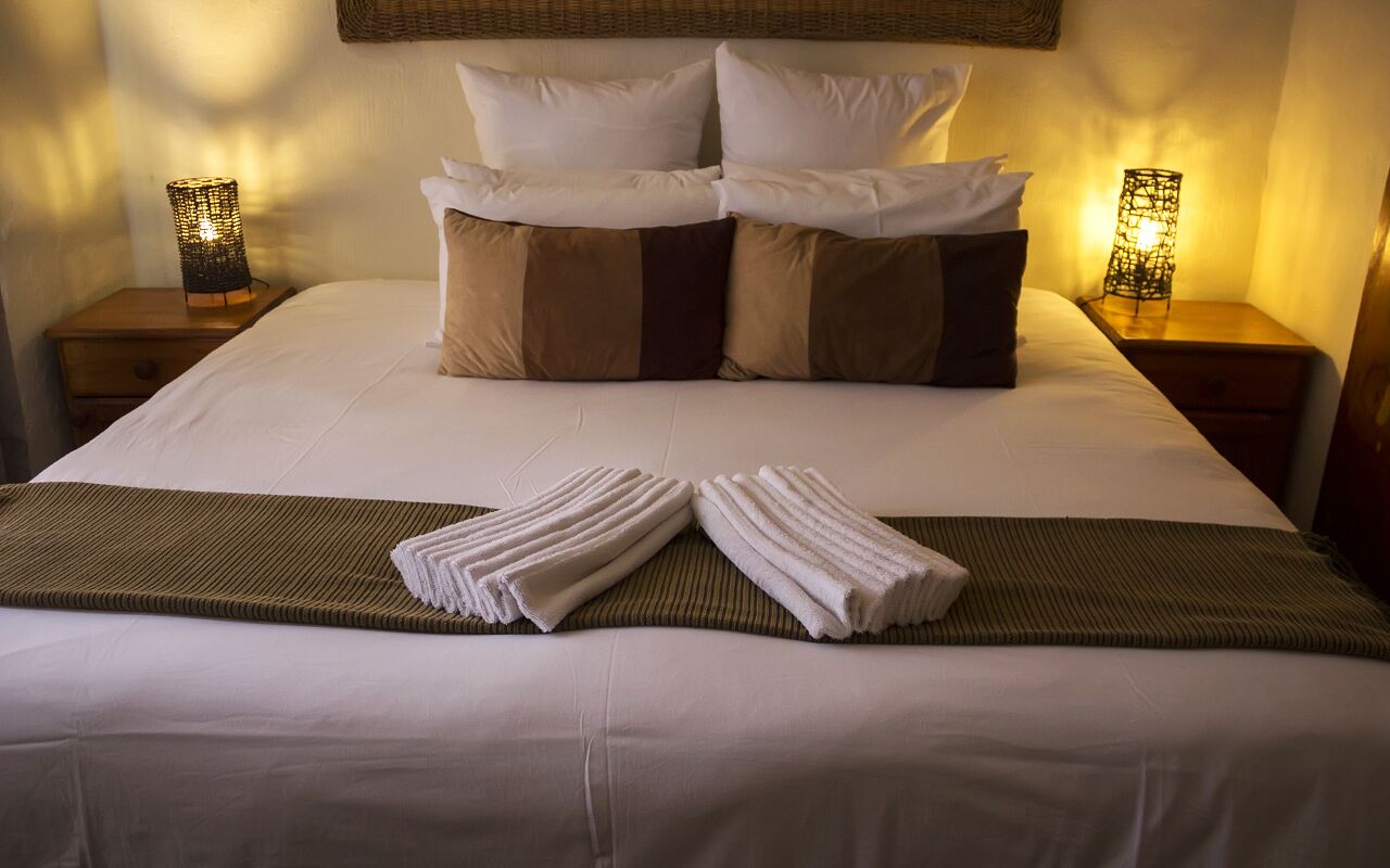 Cama bem arrumada com lençóis brancos, duas almofadas marrons e uma toalha dobrada sobre a colcha.