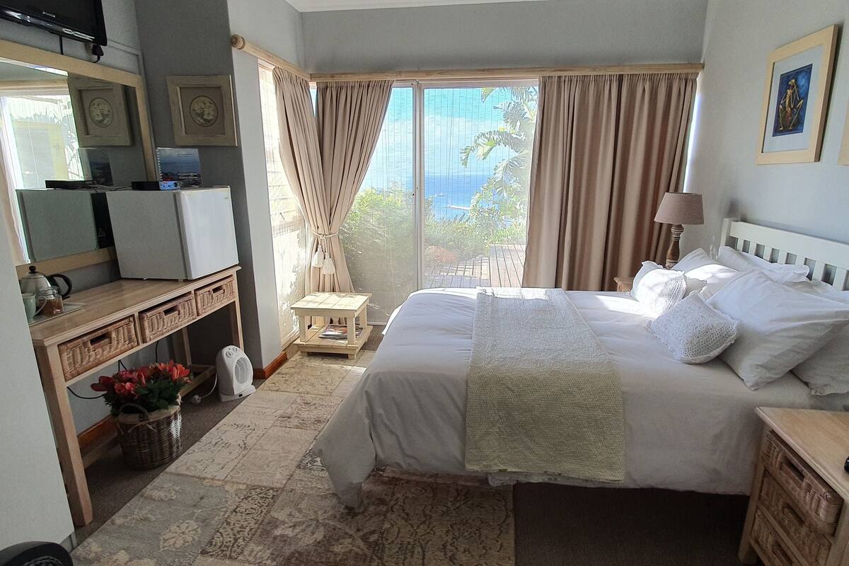 A hotel room with a bed, a bedside table with a lamp, a small table with a fan, a dresser, and a large window with curtains.