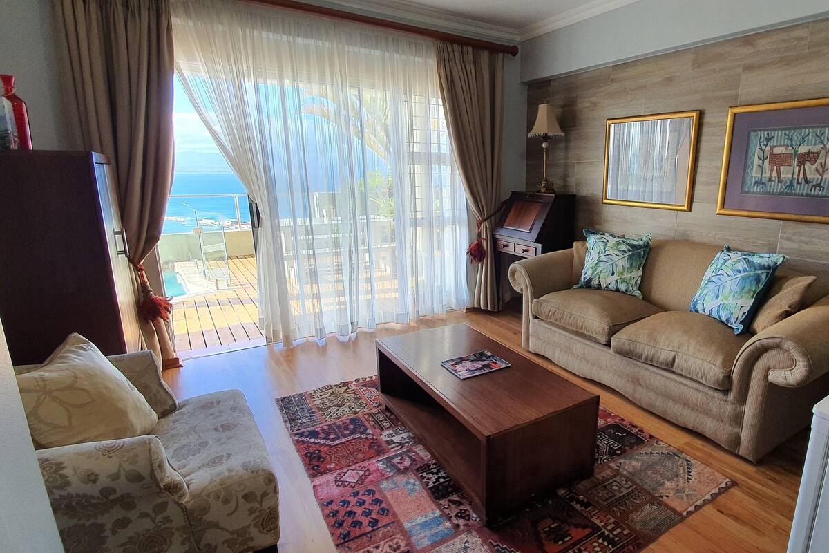 A living room with a beige sofa, patterned cushions, a wooden coffee table, and a view of the sea through sheer curtains.