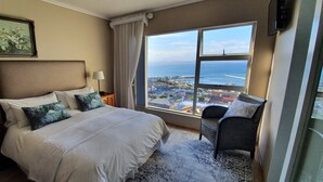 A hotel room with a large bed, two bedside lamps, a chair, and a view of the sea and cityscape. - 3 Colours Blue (Mossel Bay)