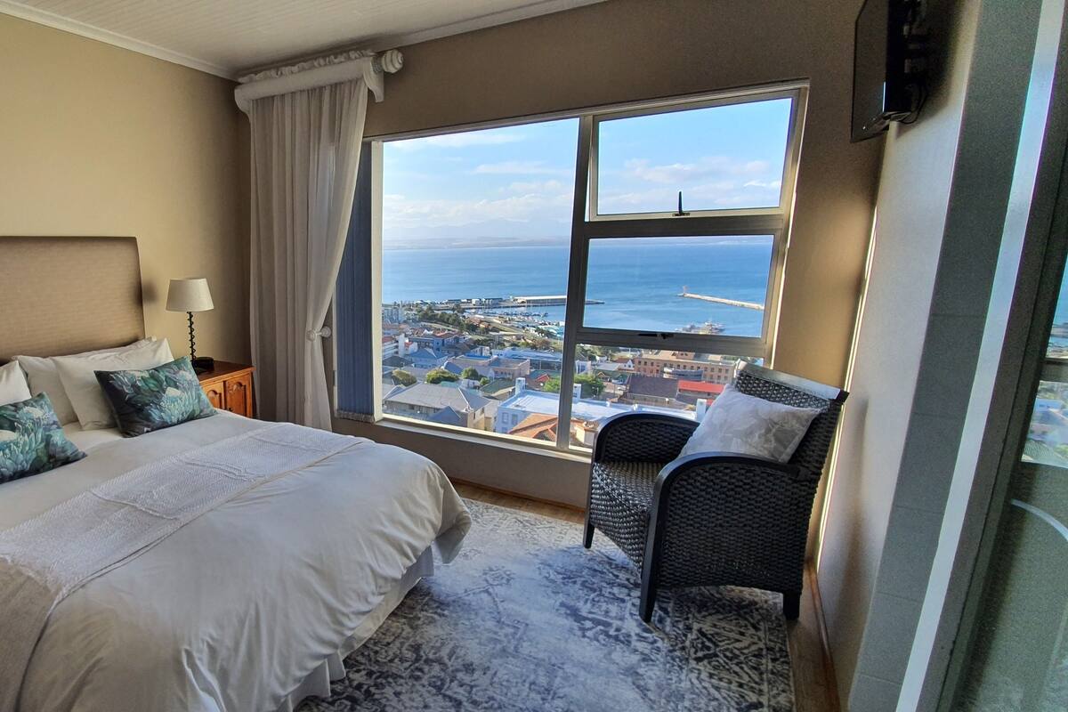 A hotel room with a large bed, two bedside lamps, a chair, and a view of the sea and cityscape.