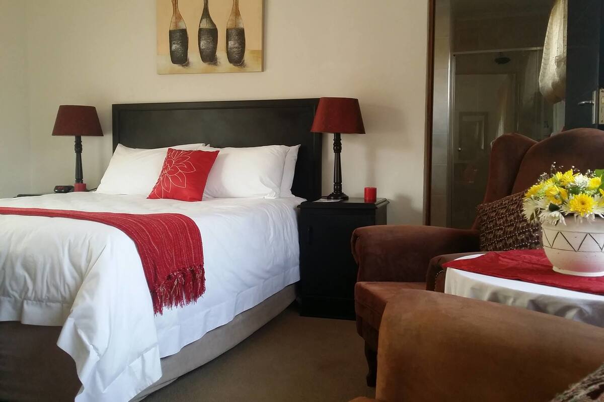 Una habitación de hotel con una cama, dos lámparas de noche, una manta roja, un cojín rojo, un jarrón con flores amarillas y un cuadro con jarrones en la pared.