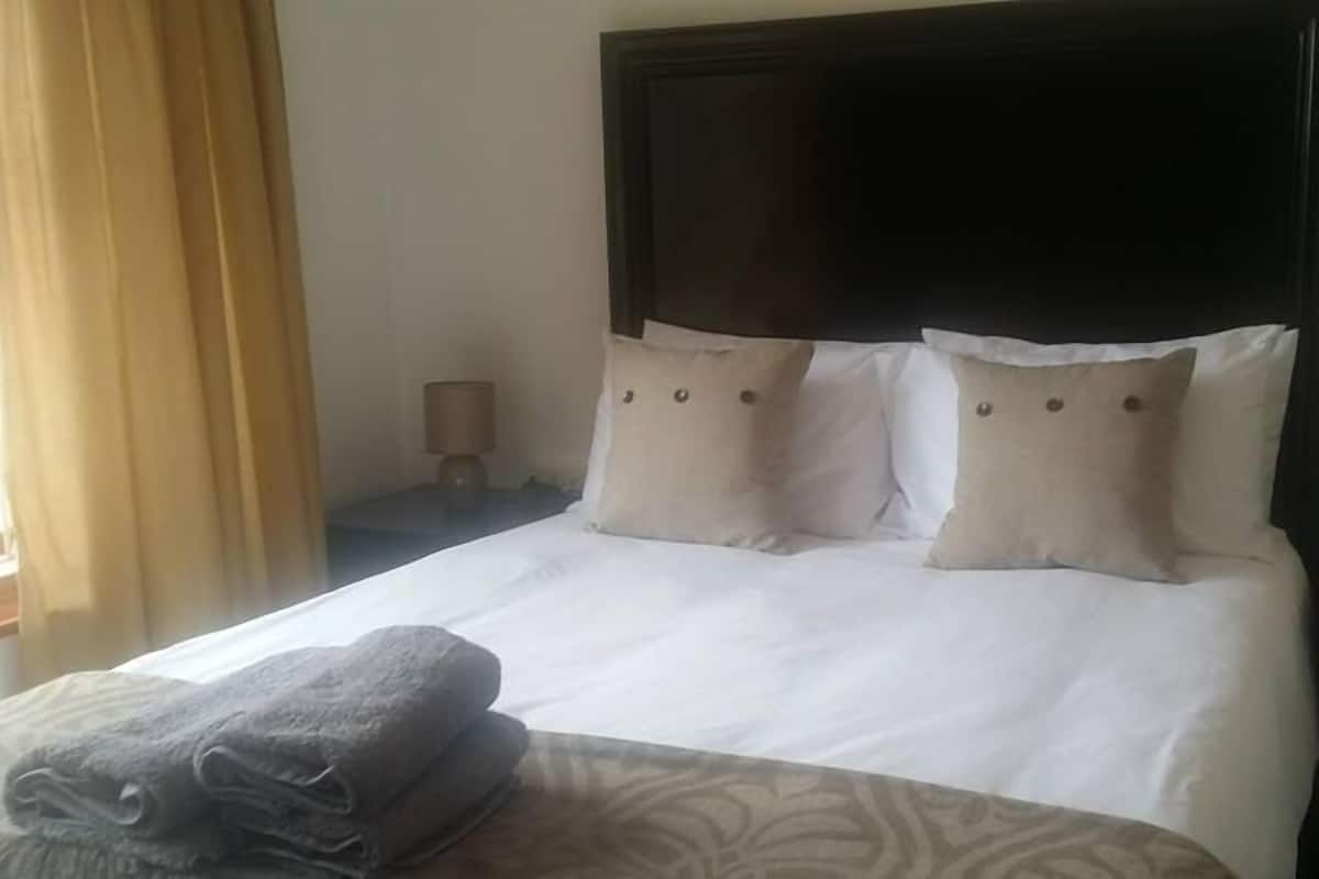 Una cama bien hecha con sábanas blancas y dos almohadas color beige, un cabecero de madera oscura y un par de toallas dobladas sobre la cama.