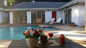 Een zwembadterras met een tafel gedekt voor drankjes, een bloemstuk, een lounge stoel en een huis met pannendak en grote ramen.