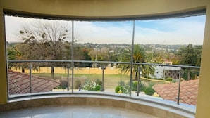 A balcony with a curved glass railing overlooking a residential area with houses and greenery. - Bella Casa Guesthouse (Sandton)