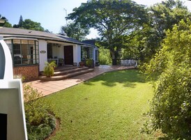 Una casa con patio frontal empedrado, un porche con sillas y un árbol grande.