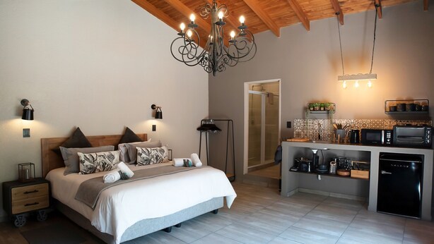 Un dormitorio moderno con una cama grande, lámparas de noche y una araña. Junto a él, una kitchenette compacta con microondas, tostadora y cafetera.