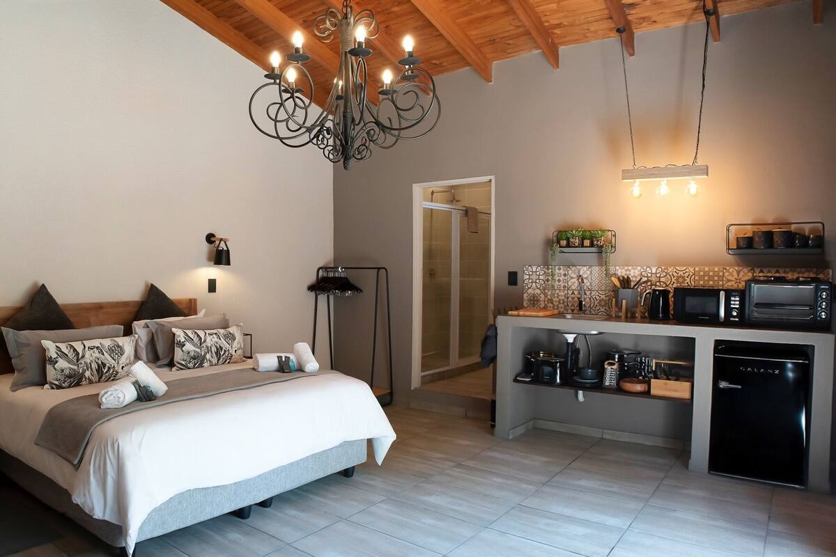 A modern bedroom with a large bed, bedside lamps, and a chandelier. Adjacent is a compact kitchenette with a microwave, toaster, and coffee maker.