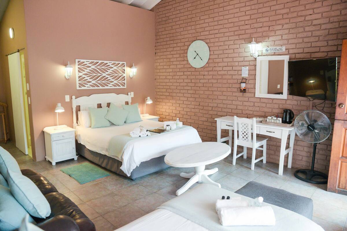 A hotel room with a bed, bedside tables, a desk, a chair, a fan, and a clock on the wall.