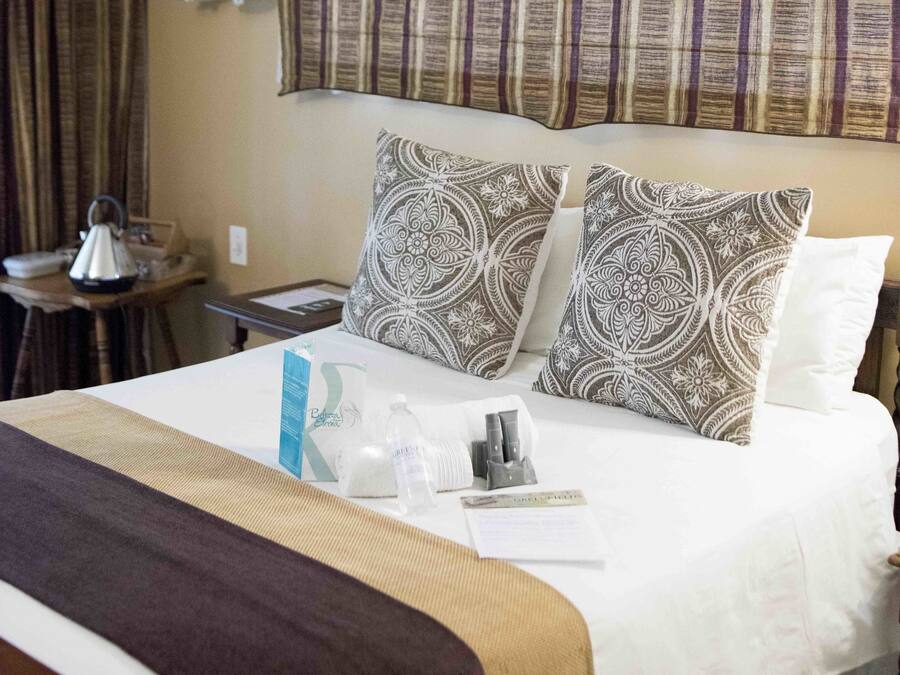 Habitación de hotel con una cama grande, dos almohadas estampadas, una mesita de noche con lámpara y una ventana con cortinas a rayas.