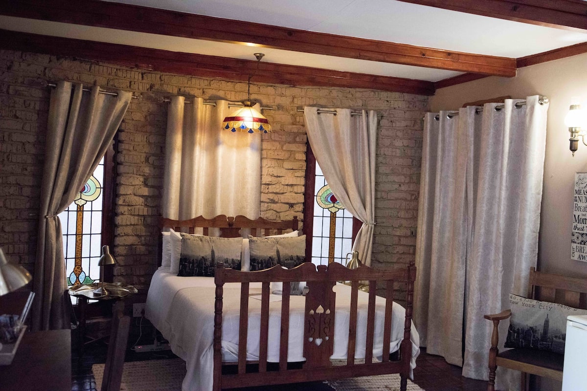A cozy room with a wooden bed, a small table with a lamp, and a window with curtains.