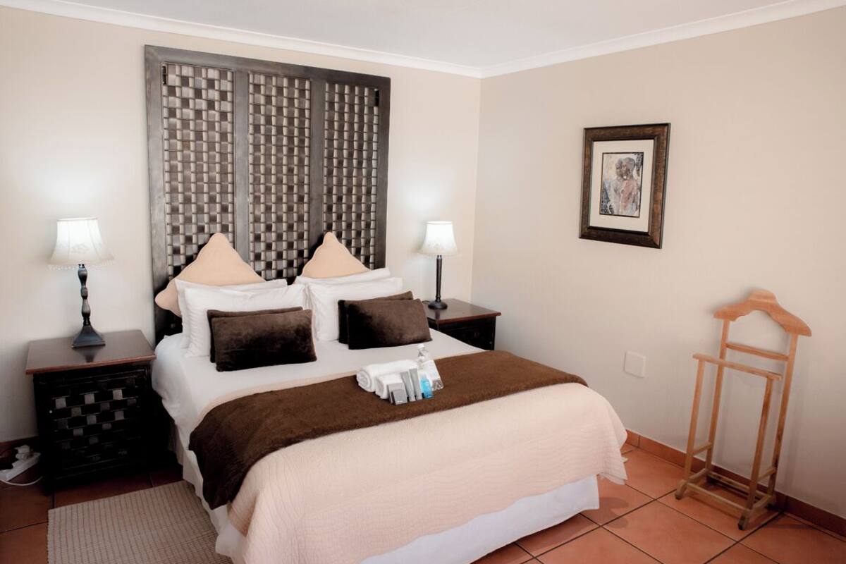 A neatly arranged hotel room with a large bed, two bedside tables, a headboard with a geometric pattern, a framed picture on the wall, and a wooden rack.