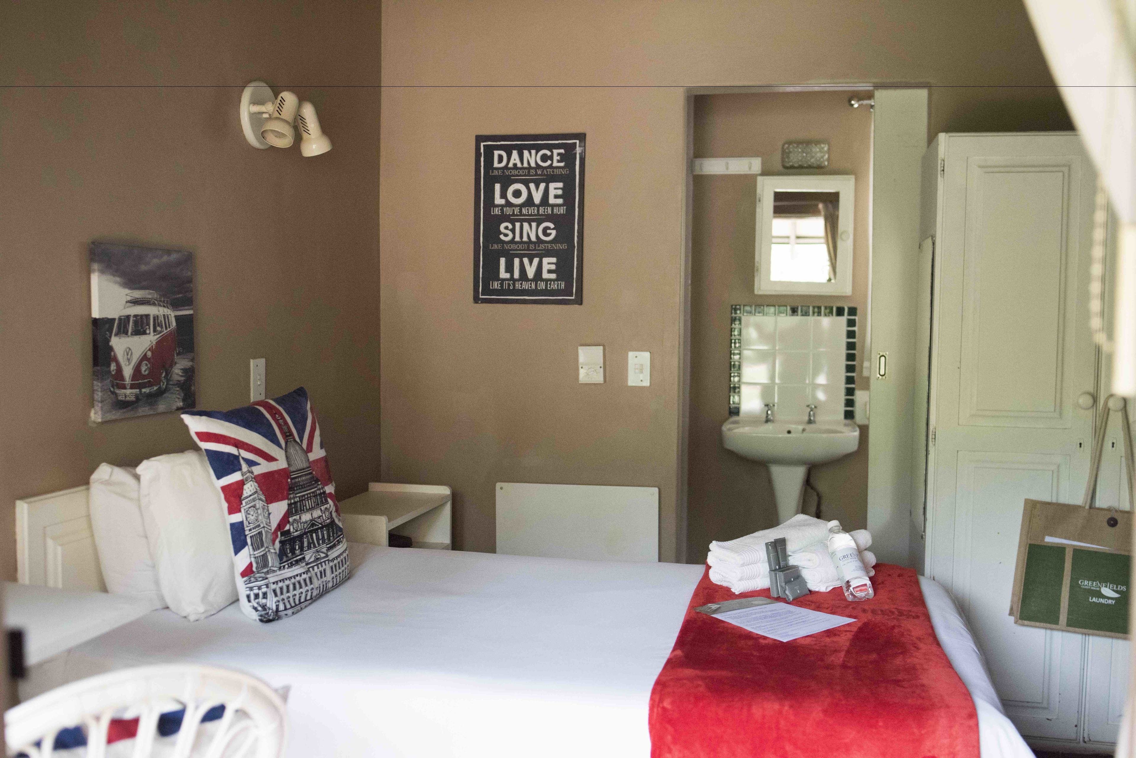 A hotel room with a bed, pillows, a red blanket, a mirror, a sink, and a sign with motivational words.