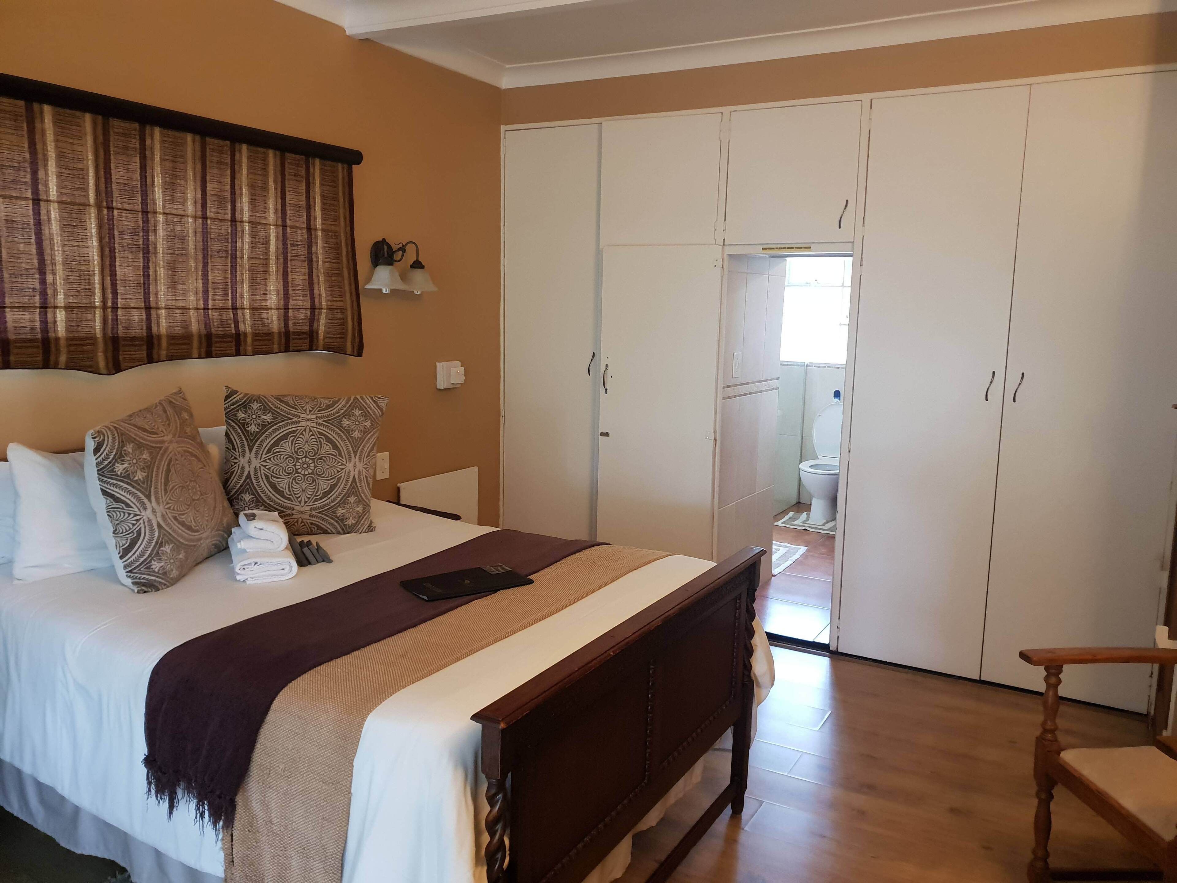 Un dormitorio con cama, almohadas, una mesita de noche, una lámpara, un armario y un baño visible a través de una puerta abierta.