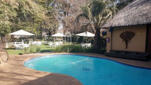 Una piscina con borde de ladrillo, rodeada de mesas y sombrillas blancas, y un edificio con techo de paja.