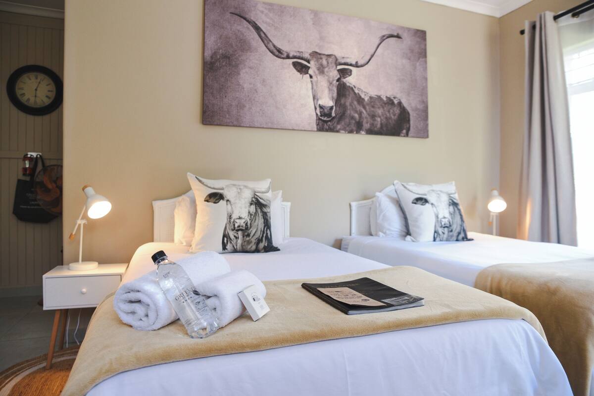 A hotel room with two beds, a clock on the wall, and a painting of a bull.