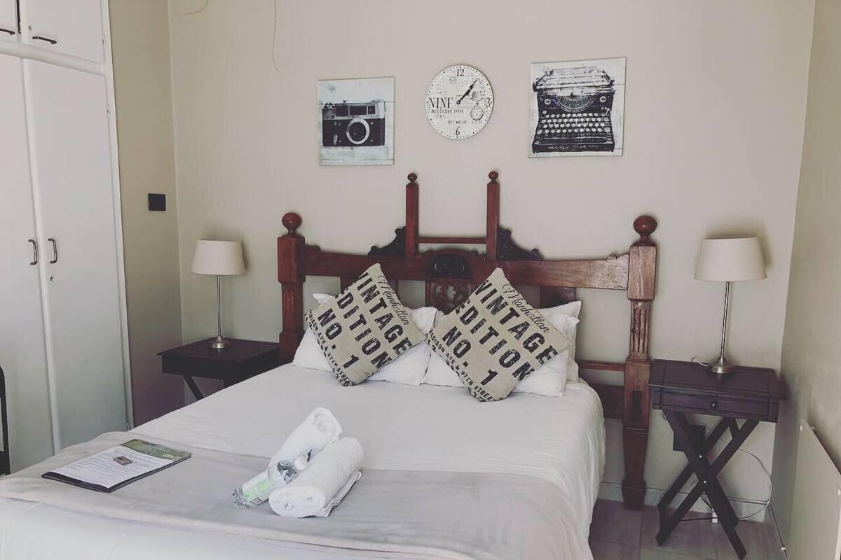 A neatly made bed with white bedding, two bedside tables with lamps, a wall clock, and vintage-themed wall art.