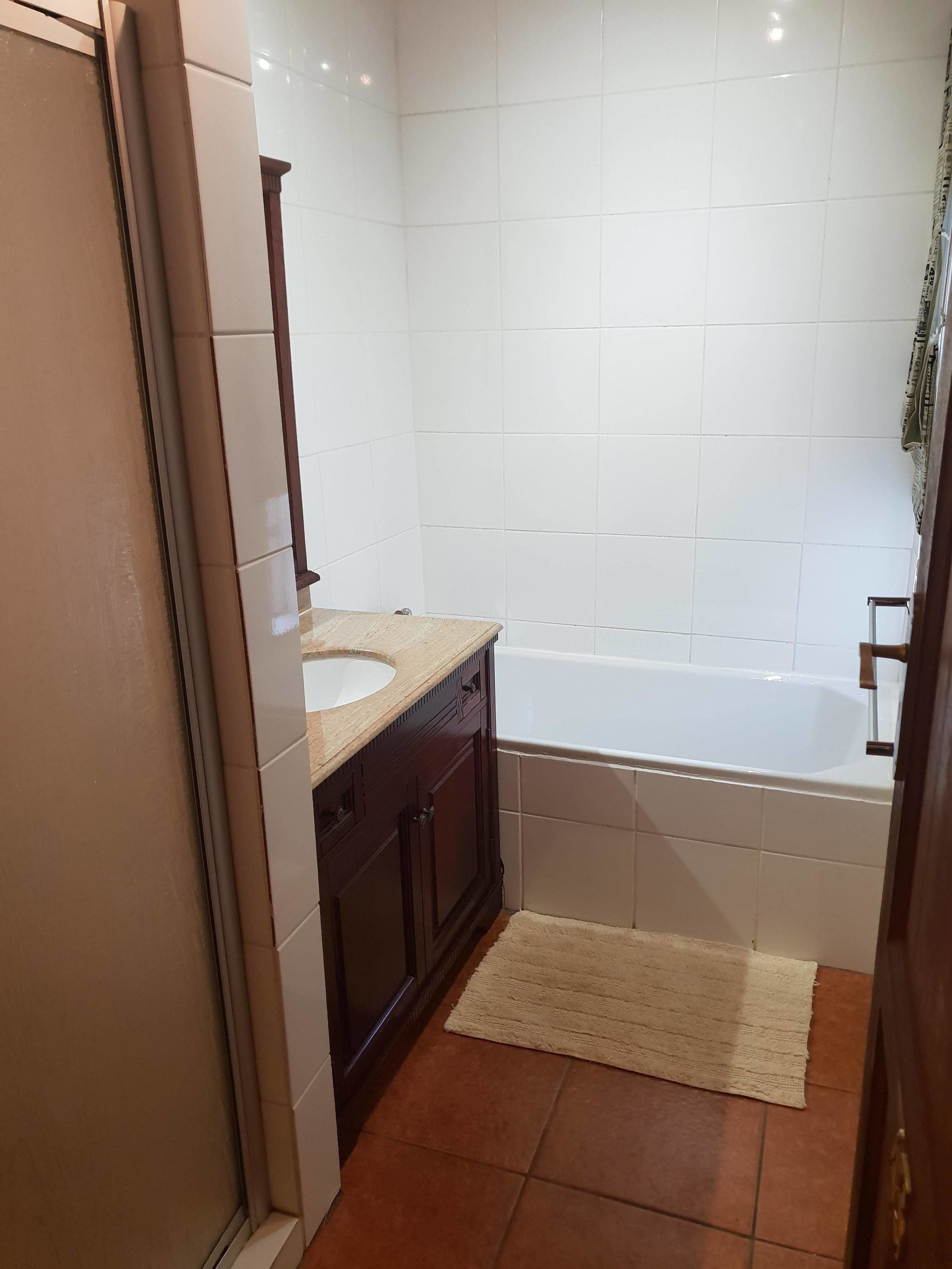 A bathroom with a white bathtub, a wooden vanity with a sink, and a beige rug on the floor.