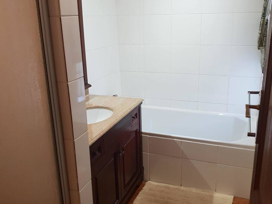 A bathroom with a white bathtub, a wooden vanity with a sink, and a beige rug on the floor.