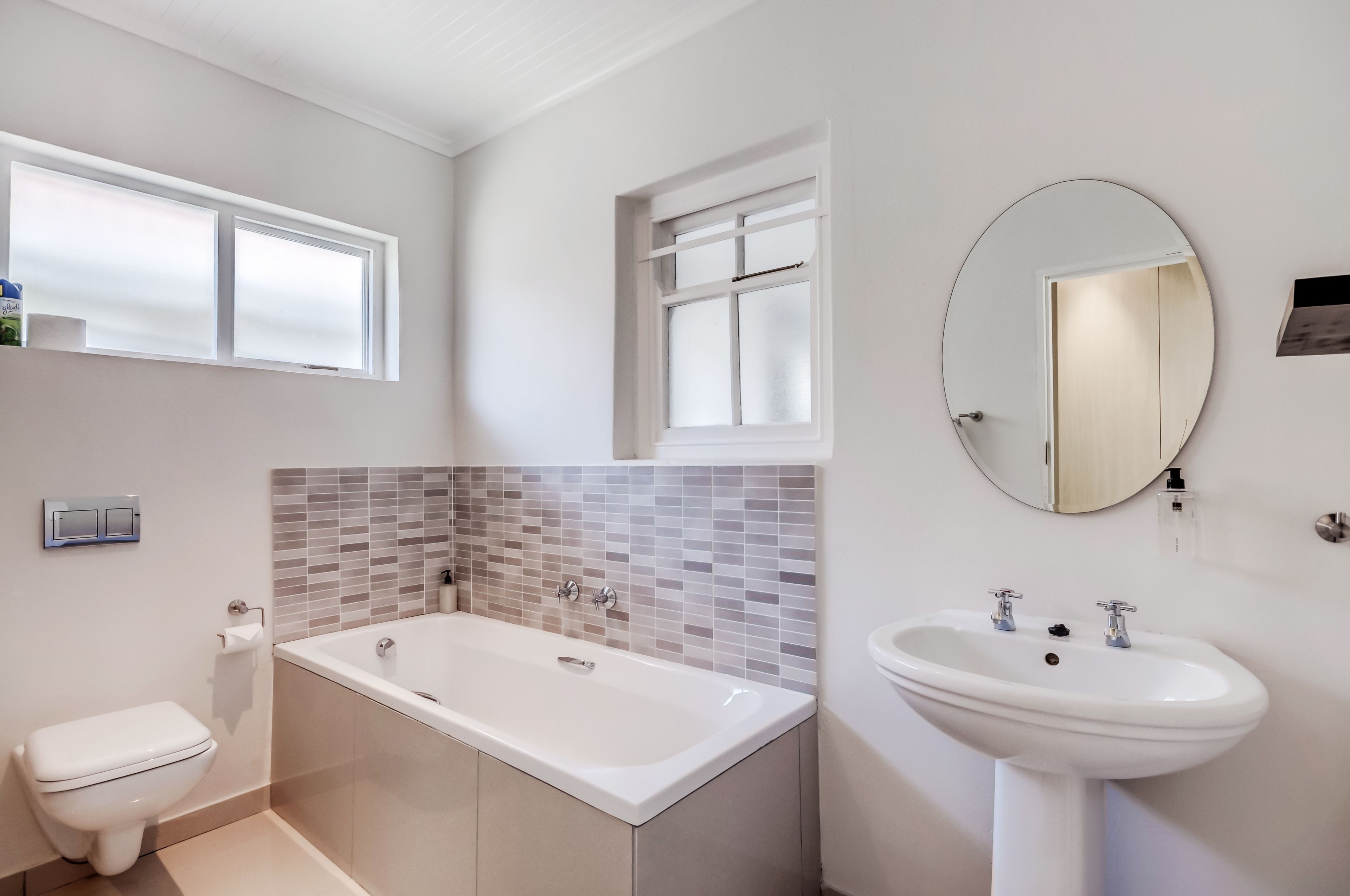 A modern bathroom with a white bathtub, a sink, and a toilet. There is a window above the bathtub and a mirror on the wall.