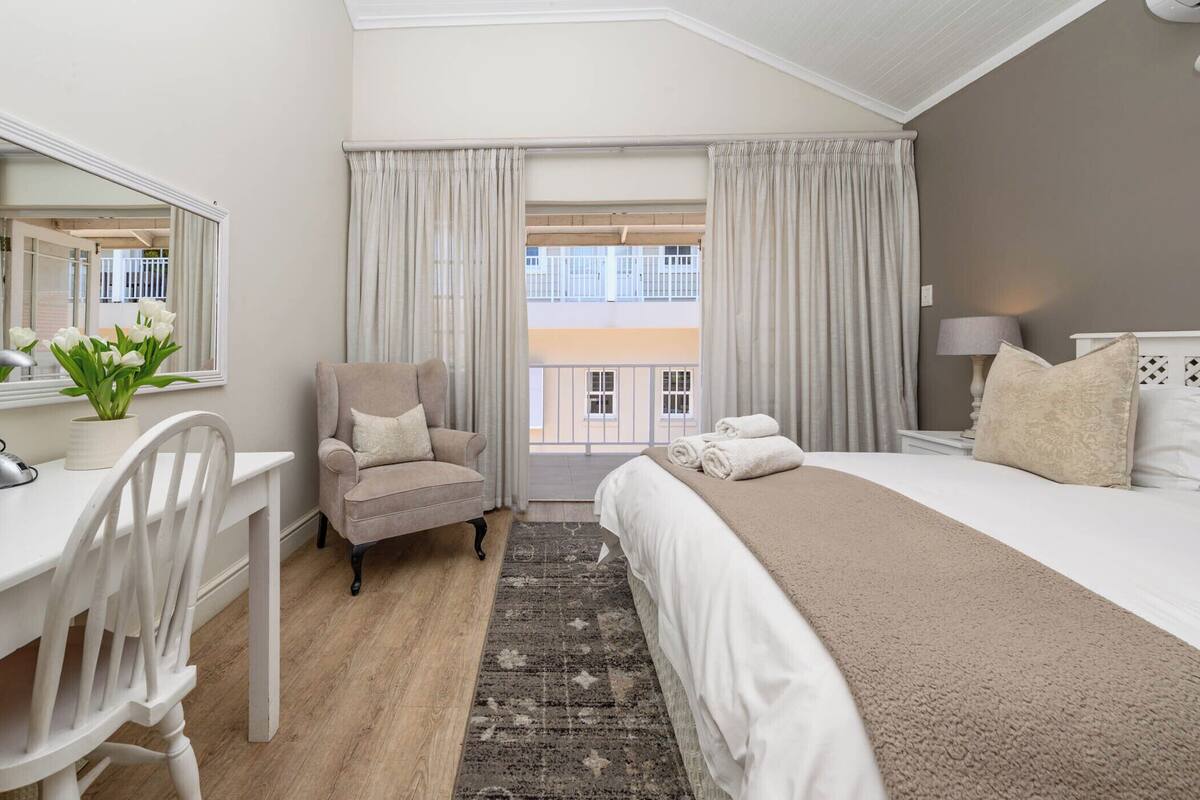 Habitación de hotel con cama, escritorio, silla, ventana grande con cortinas y vista a edificios.