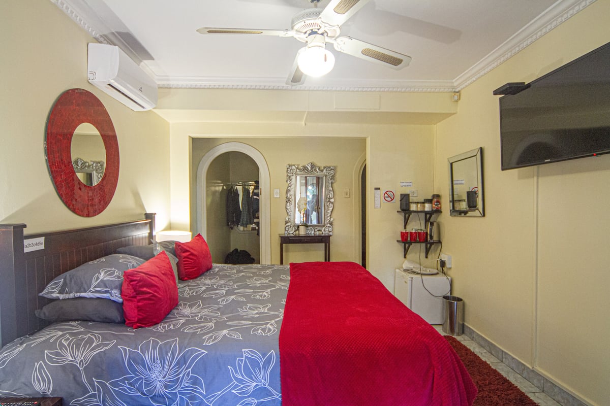 A hotel room with a bed, a ceiling fan, a TV, a mirror, and a door leading to a bathroom.
