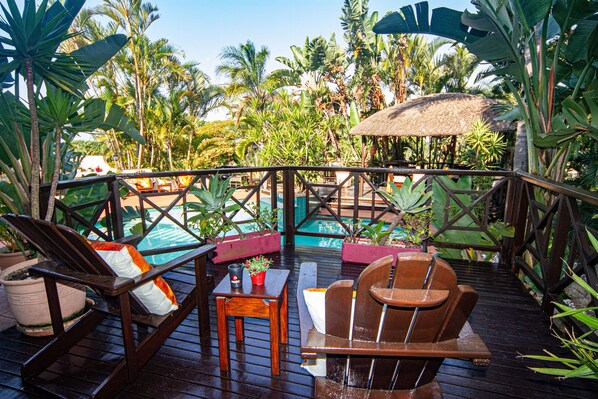 A wooden deck with lounge chairs and a table, overlooking a pool area surrounded by tropical plants. - La Loggia B&B (Umhlanga)