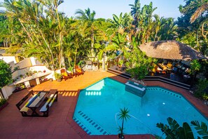 A swimming pool surrounded by lush greenery, with a thatched umbrella and wooden deck chairs. - La Loggia B&B (Umhlanga)