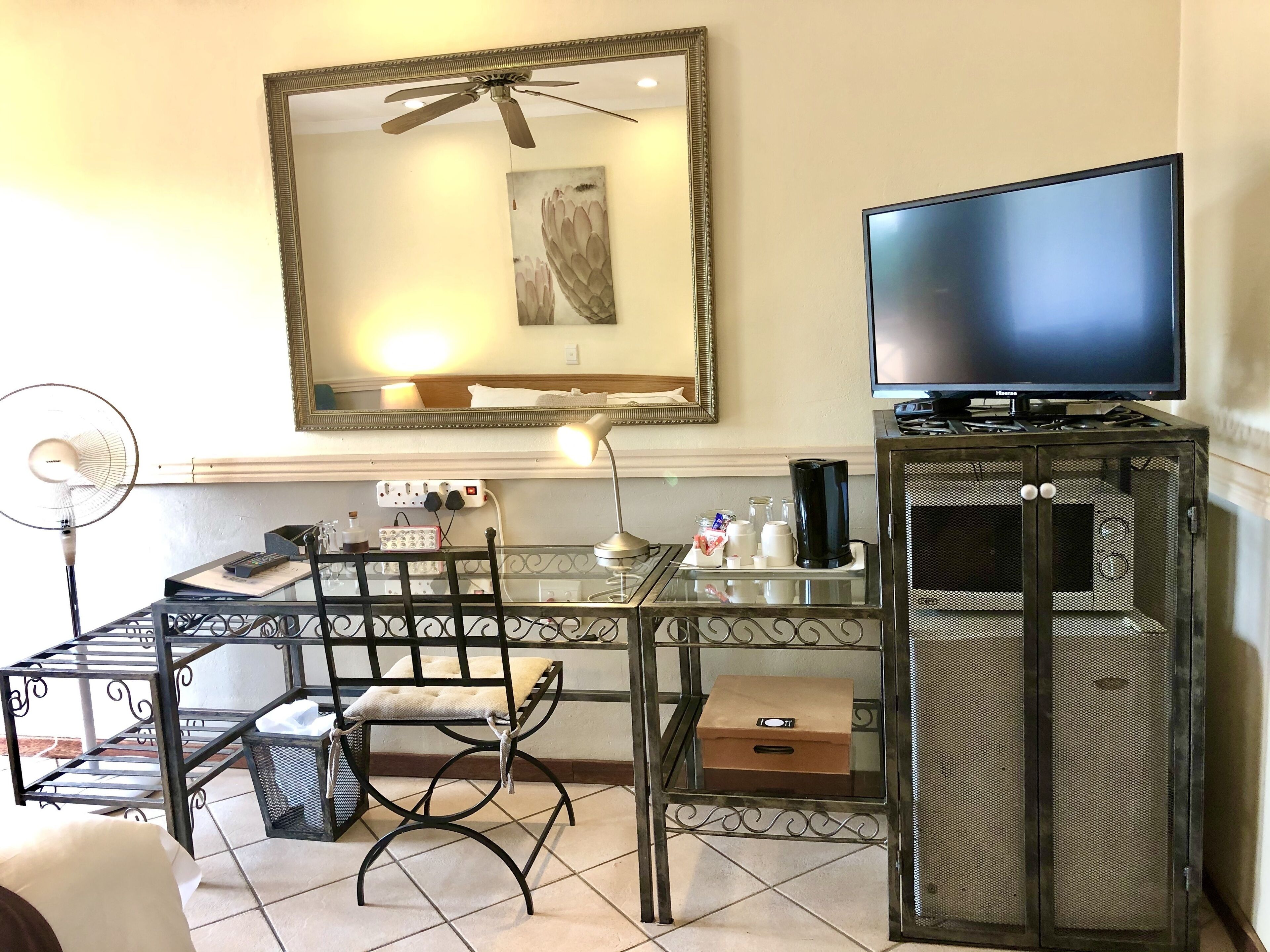 Ein Hotelzimmer mit Bett, Fernseher, Schreibtisch mit Stuhl, Ventilator und einem kleinen Tisch mit Lampe.