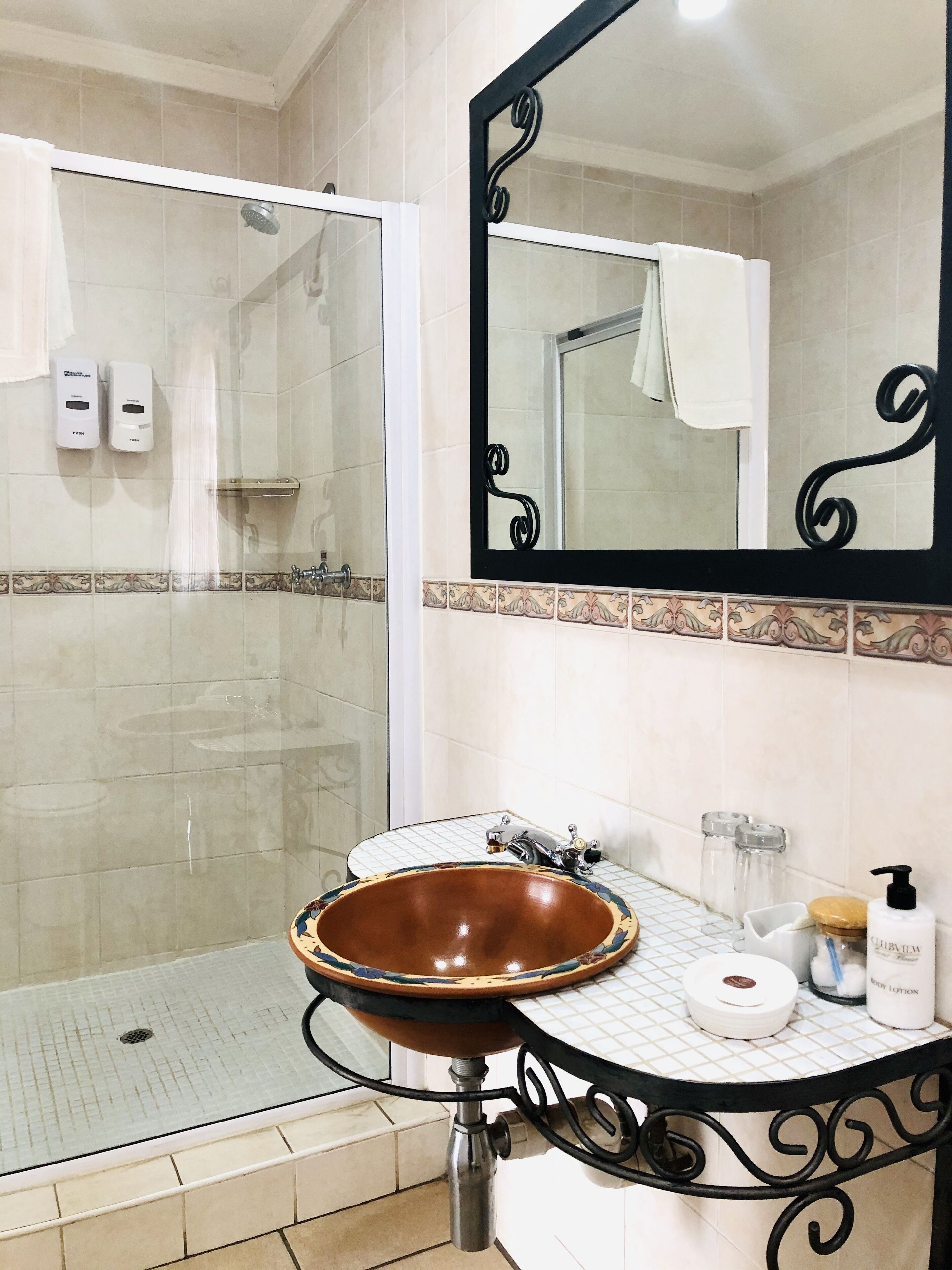 A bathroom with a glass shower, a brown ceramic sink, a mirror, and toiletries on the counter.
