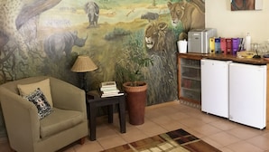 A living room with a mural of a savannah scene, a sofa, a small table with books, a lamp, a potted plant, and a kitchenette area.