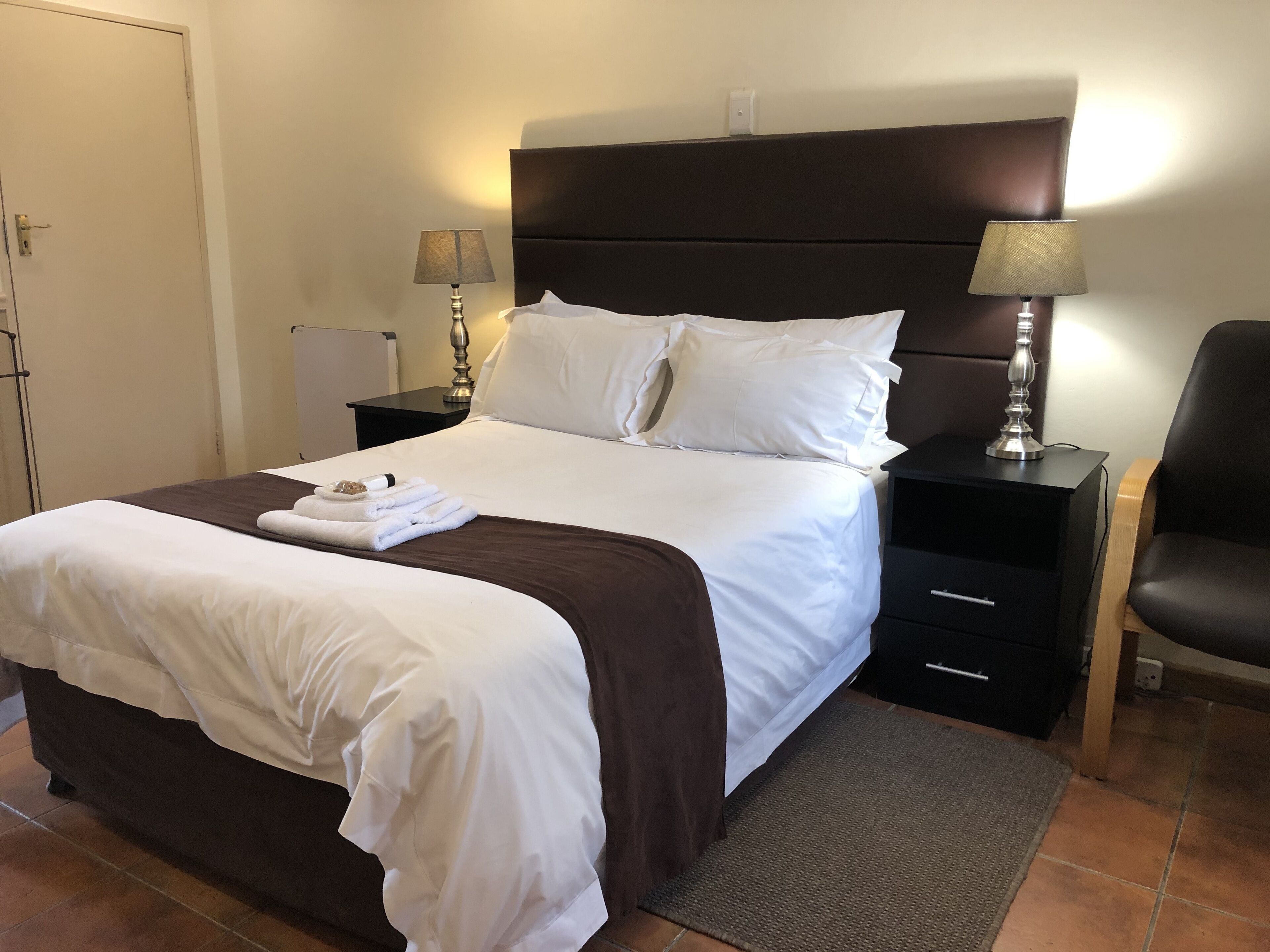 A neatly made bed with white linens and a brown accent, two bedside tables with lamps, a chair, and a door.