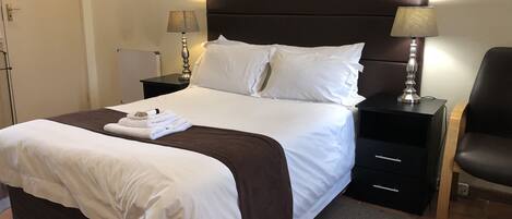 A neatly made bed with white linens and a brown accent, two bedside tables with lamps, a chair, and a door.
