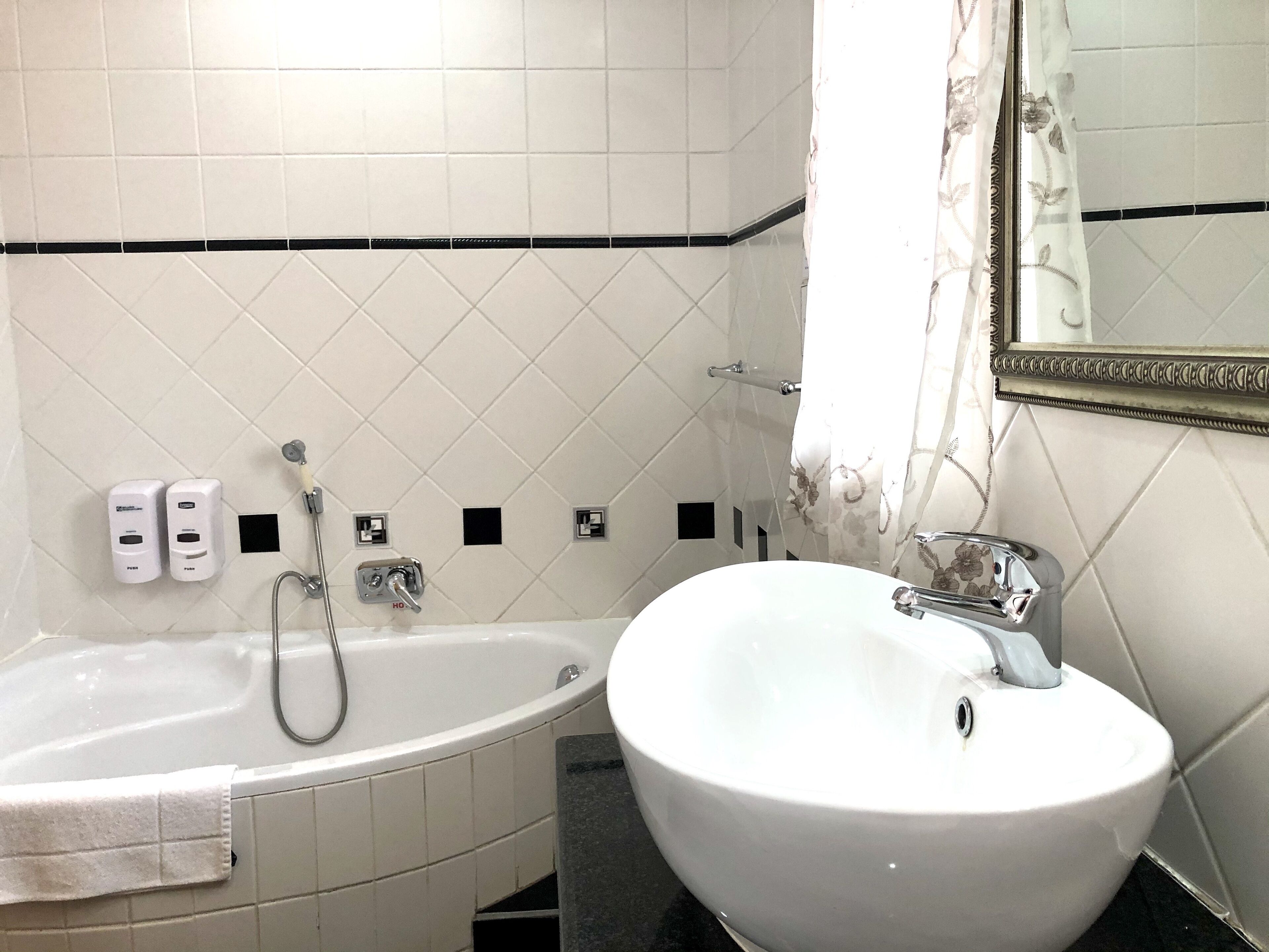 Un baño con una bañera blanca, un lavamanos blanco y un espejo. Hay dos dispensadores de jabón en la pared y una cortina de ducha con estampado floral.