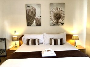 A hotel room with a large bed, two pillows, two bedside lamps, and two wall-mounted artworks. - Clubview Guest House (Centurion)