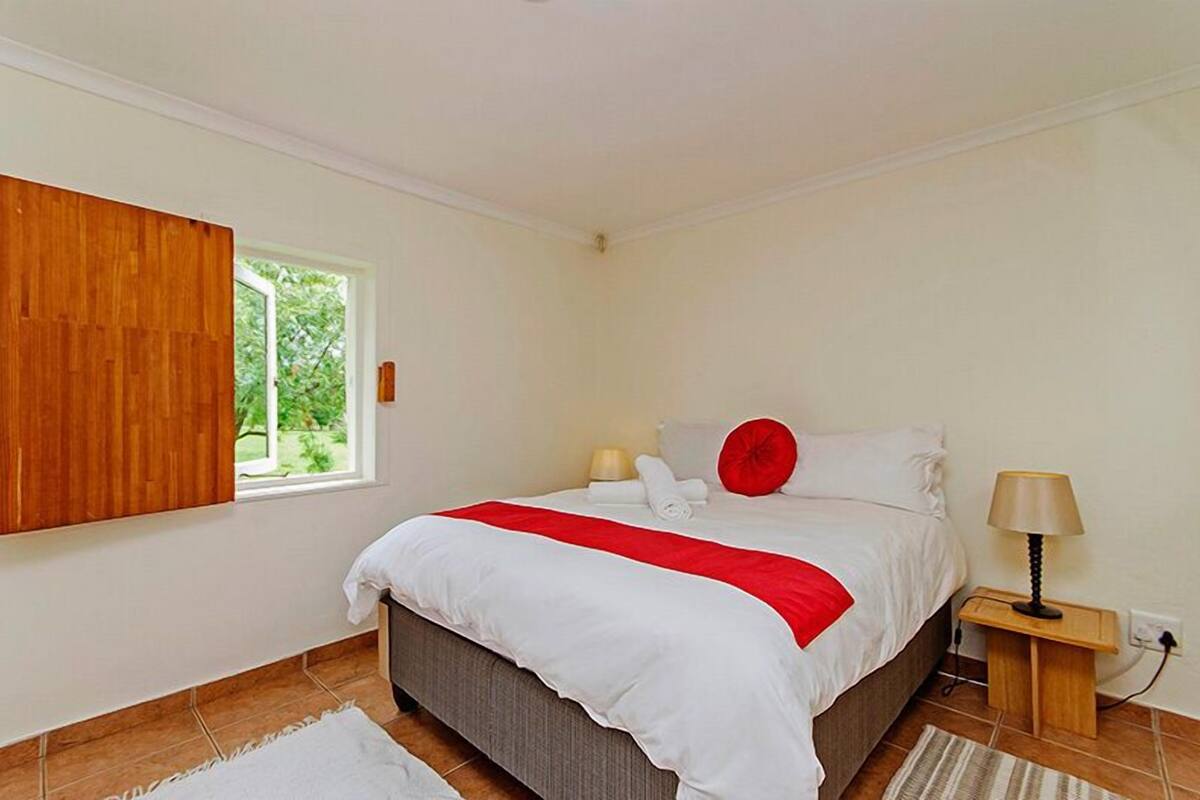 Ein Schlafzimmer mit einem Bett, einem Nachttisch, einer Lampe, einem Fenster mit Vorhängen und Blick ins Grüne.