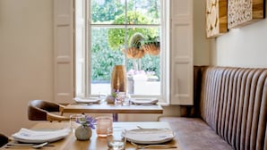 A dining area with a wooden table set for two, a cushioned bench, and a window with a view of greenery outside. - Macaron Boutique Guest House (Franschhoek)