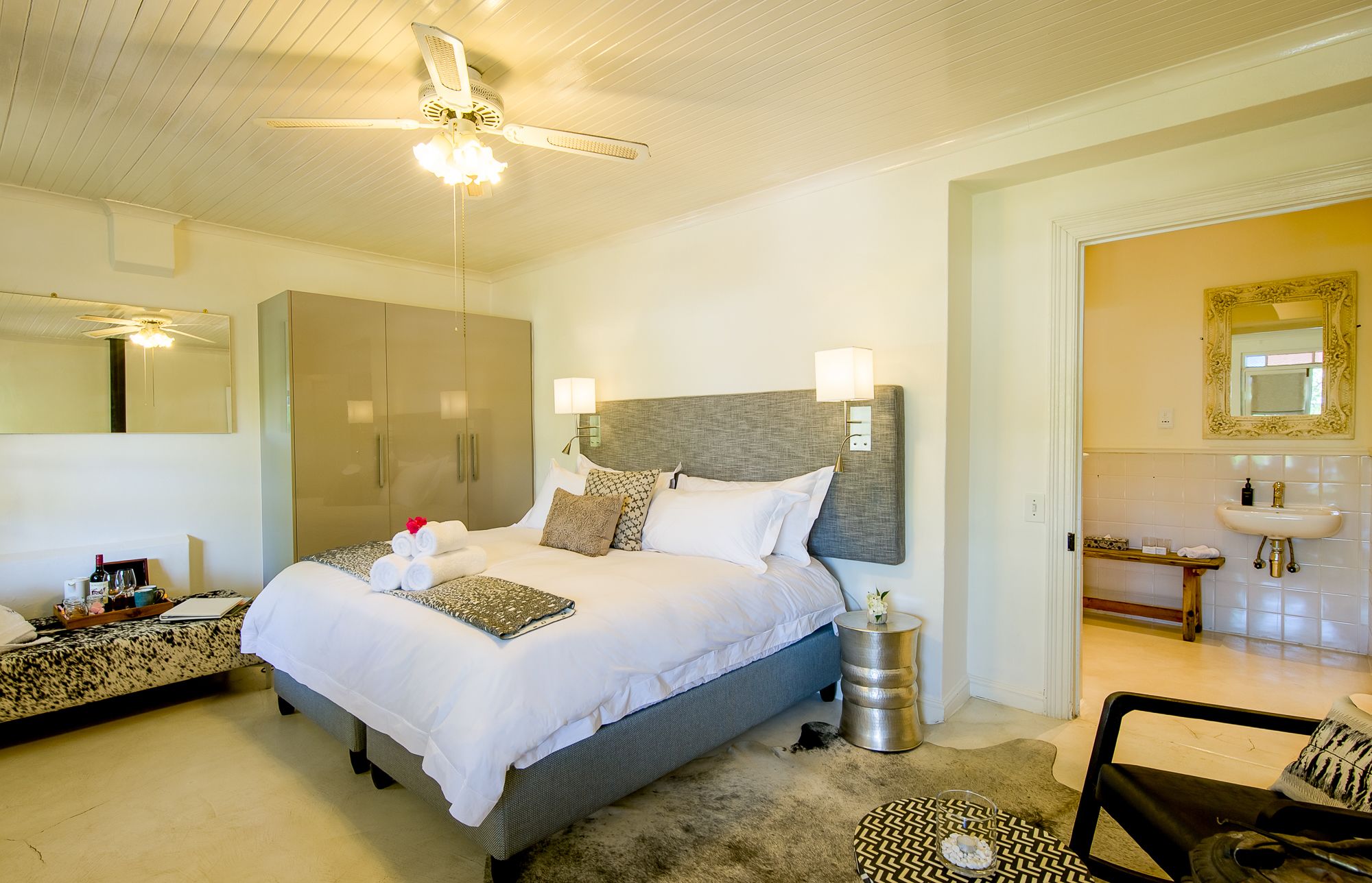 A well-lit bedroom with a large bed, bedside tables, a wardrobe, and a bathroom visible through an open door.