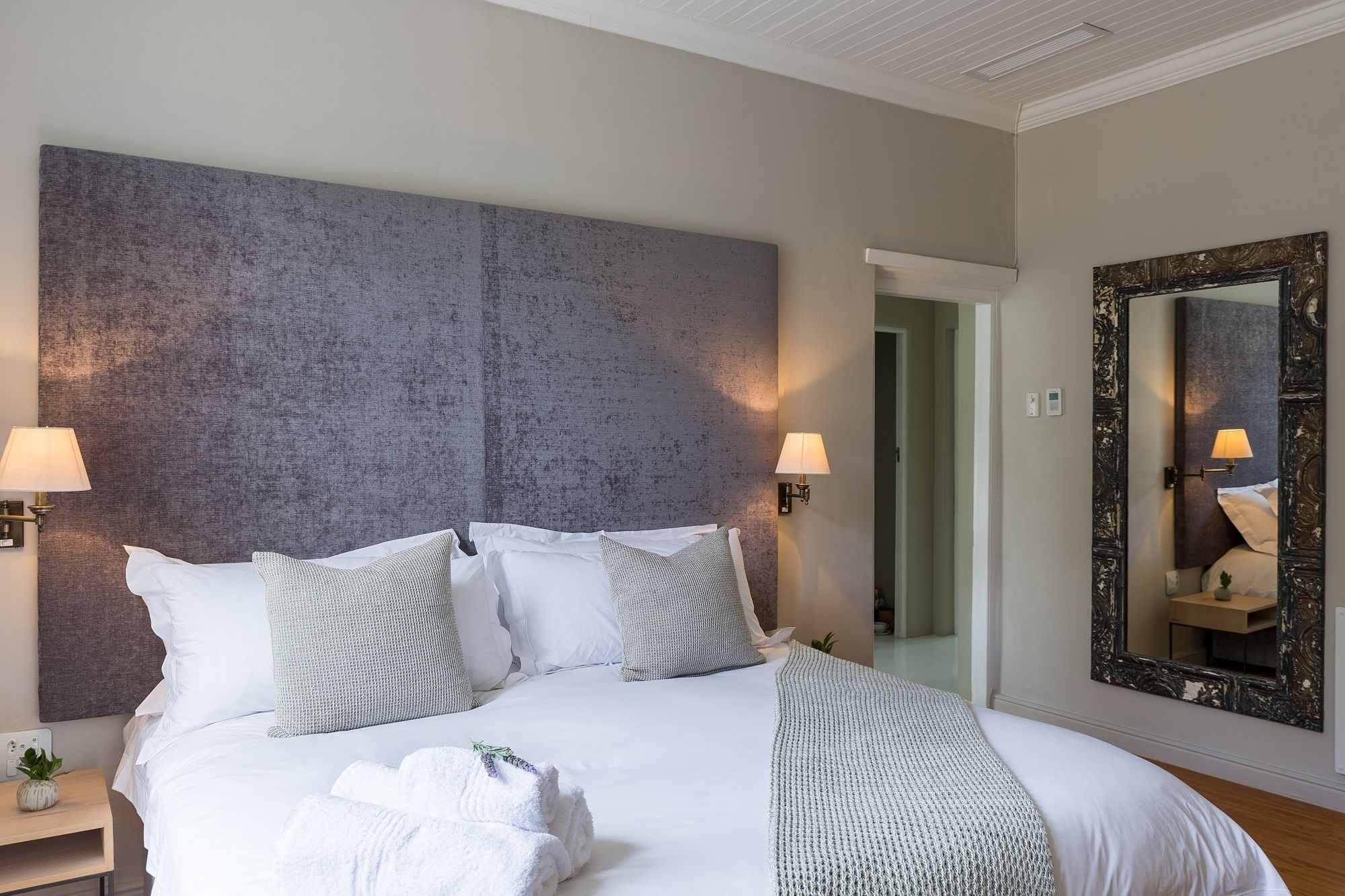 Una cama bien hecha con sábanas blancas y varias almohadas, una mesita de noche con lámpara y un espejo grande en la pared.