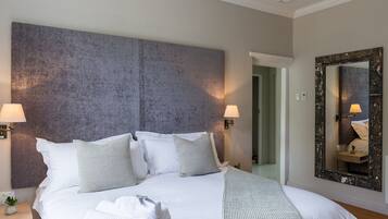 Una cama bien hecha con sábanas blancas y varias almohadas, una mesita de noche con lámpara y un espejo grande en la pared.
